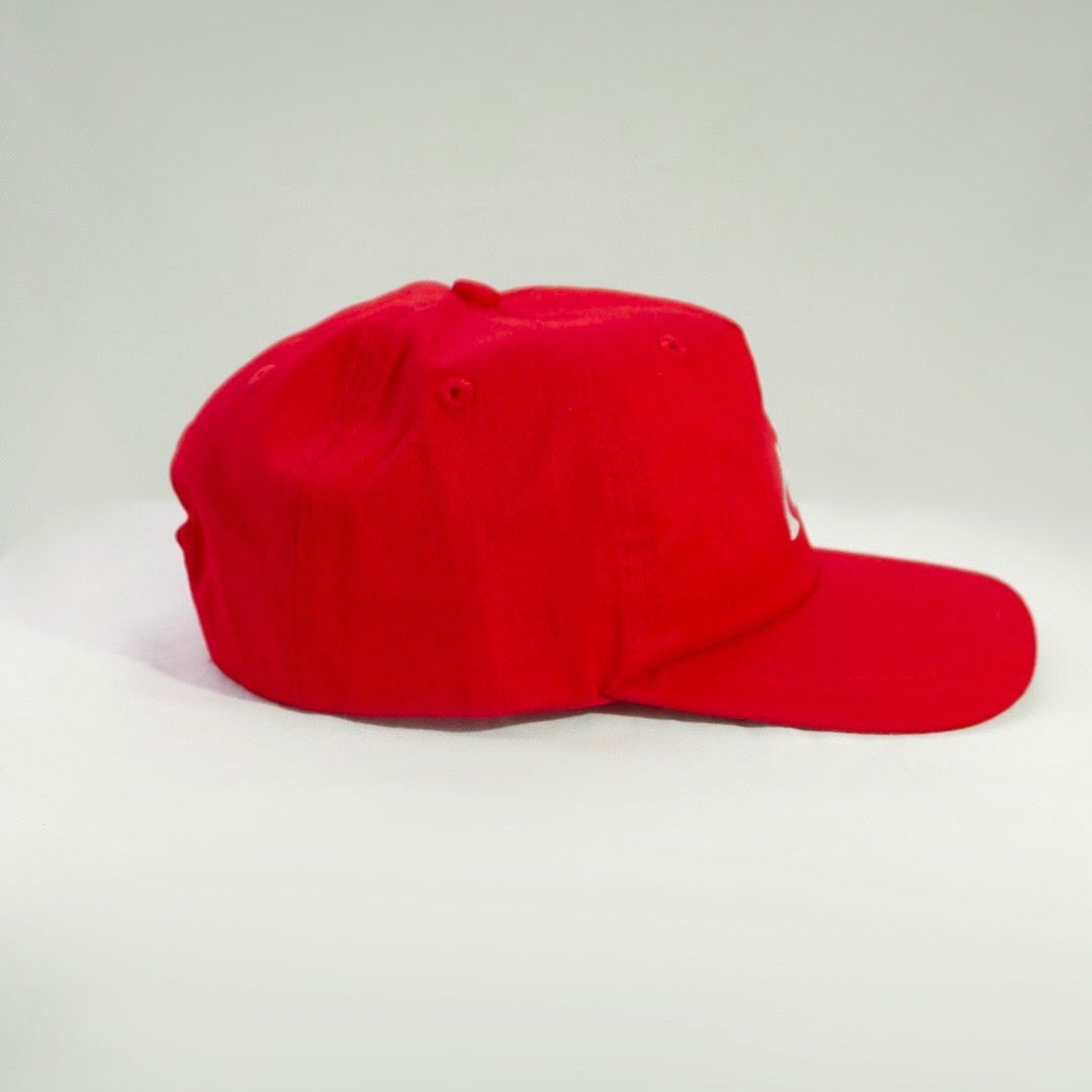A bright red baseball cap with a curved brim, photographed from the side against a neutral light gray studio background.