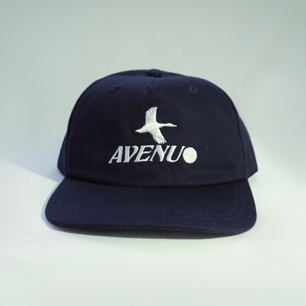 A dark navy blue snapback cap with a white embroidered seagull logo and the word 'AVENU' on the front, set against a light gray gradient background.