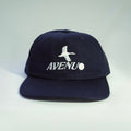 A dark navy blue snapback cap with a white embroidered seagull logo and the word 'AVENU' on the front, set against a light gray gradient background.