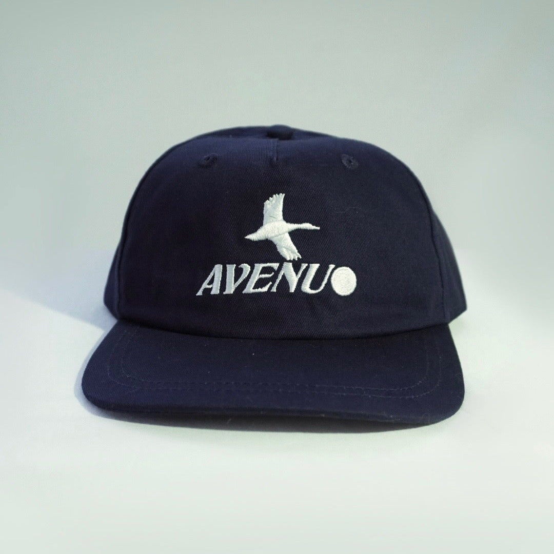 A dark navy blue snapback cap with a white embroidered seagull logo and the word 'AVENU' on the front, set against a light gray gradient background.