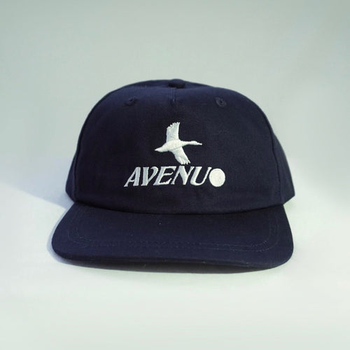 A dark navy blue snapback cap with a white embroidered seagull logo and the word 'AVENU' on the front, set against a light gray gradient background.