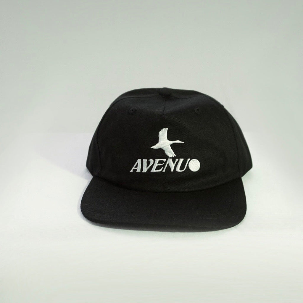 A black flat-brim cap with a white embroidered logo featuring a bird silhouette and the text 'AVENU' on the front, set against a light gray gradient background.